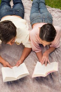 Man and a woman reading books while lying prone on a grey blanket in the grass