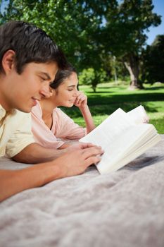 Man and a woman reading books while lying prone on a grey blanket int the grass with trees in the background