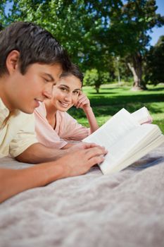 Woman looking to the side as she reads a book with her friend while lying prone on a grey blanket in the grass with trees in the background