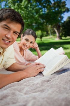 Two friends looking towards the side while reading books while lying on a grey blanket in the grass with tress in the background