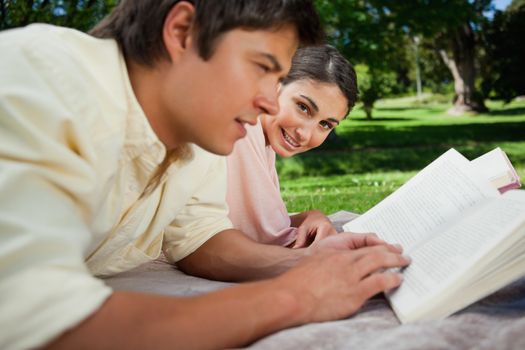 Woman looks towards the side while reading with her friend as they lie prone on a blanket in the grass with trees in the background