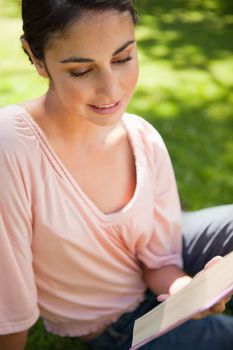 Woman smiling while she reads a book as she sits down on grass on a sunny day