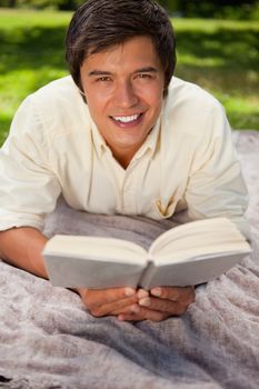 Man looking straight ahead smiling while reading a book as he lies on a grey blanket in the grass