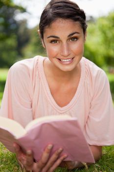 Woman looking ahead and smiling while reading a book as she is lies in grass with trees in the background