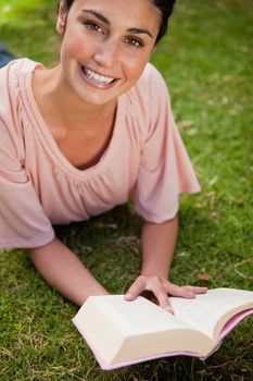 Smiling woman looking upwards while reading a book as she is lying prone on grass