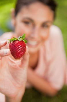 Woman holds a strawberry at arms reach while lying prone in grass with focus on the stawberry