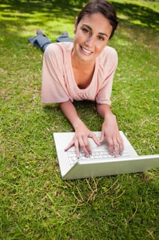 Woman looking straight ahead while she uses a laptop as she is lying down in grass