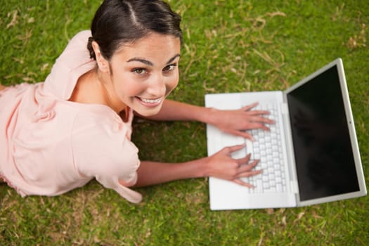 Woman looking upwards while she uses a laptop as she is lying prone in grass