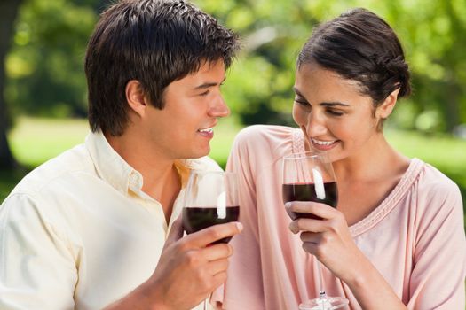 Man and a woman holding glasses of red wine while smiling in a park with trees in the background