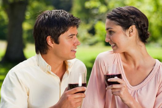 Man and a woman smiling st each other while each  holding a glass of red wine in a park