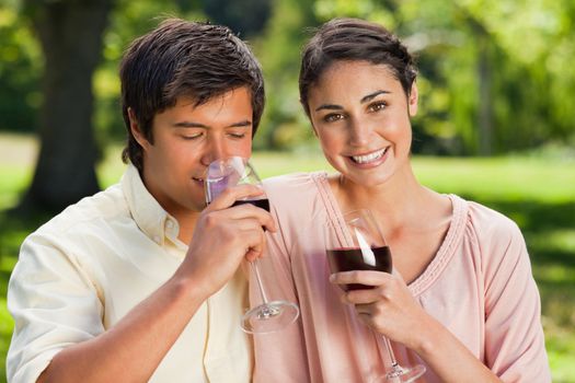 Woman looking ahead and smiling while her friend drinks from a glass of red wine in a park