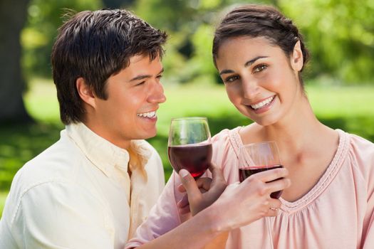 Woman looks ahead while smiling as she is linking arms with her friend and holding glasses of red wine in a park