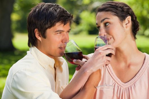 Man and a woman drinking wine while linking their arms together in a park