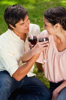 Two friends smiling while they look at each other as they are linking their arms and holding glasses of wine in a park