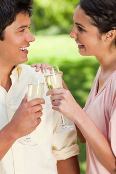 Man and a woman smiling while looking at each other as they touch glasses of champagne in a park