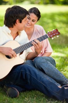 Woman smiling while looking at her friend play the guitar as they sit on grass togethe
