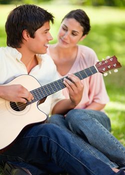 Man looking ahead and smiling as he is playing the guitar and being watched by his friend who is sitting next to him on the grass