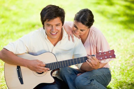 Woman putting her hand on her smiling friends shoulder while he plays the guitar as they both sit on the grass