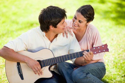Man plays the guitar while looking at his friend who has her hand on his shoulder as they both sit on the grass