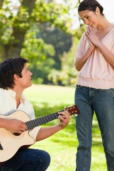 Woman standing and smiling with her hands joined as she admires her friend who is playing the guitar