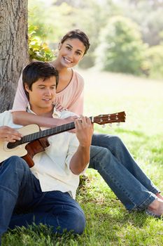 Woman sitting behind her friend and resting her arm on his chest while he plays a guitar under a tree