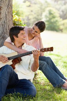 Woman looking at her friend and resting her arm on his chest while he plays a guitar under a tree