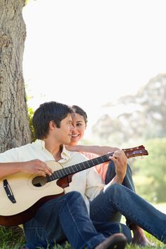 Man sitting against the trunk of a tree while playing a guitar as his friend watches him