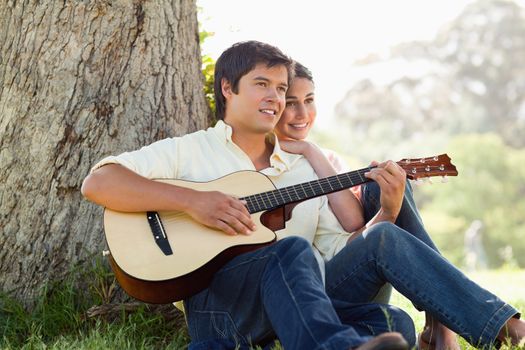 Man and his friend look into the distance as they listen to him playing the guitar while sitting against the trunk of a tree