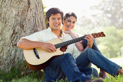 Man and his friend look straight ahead as they listen to him playing the guitar while sitting against the trunk of a tree