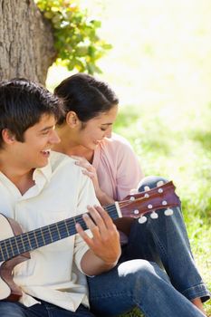 Woman laughing while leaning against her friend who is playing the guitar as they both against the trunk of a tree