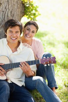 Woman smiling while resting her hand on the shoulder of her friend who i holding a guitar as they both sit against a tree