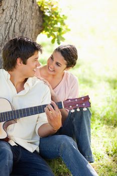 Smiling woman looking eye to eye with her friend who is holding a guitar as they both sit against a tree