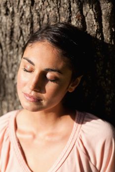 Woman sitting against the trunk of a tree with her eyes closed
