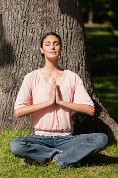 Woman with her eyes closed performing a yoga pose with her hands joined in front of her while sitting near the base of a tree