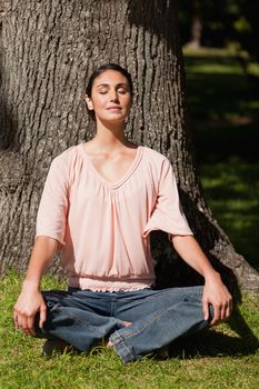 Woman with her eyes closed performing a yoga pose with her hands placed on her knees while sitting near the base of a tree