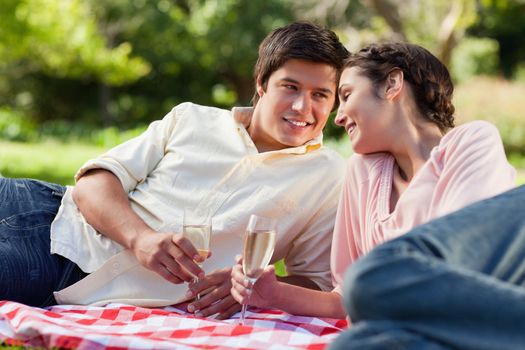 Man smiling as he looks at his friend while they touch glasses of champagne and lie down on a picnic blanket