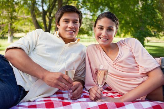 Man and a woman smiling and holding glasses of champagne as they lie down on a picnic blanket 
