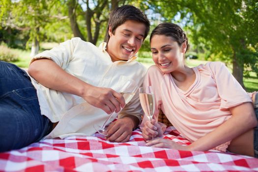 Two friends smiling while looking at glasses of champagne while lying on a red and white blanket during a picnic