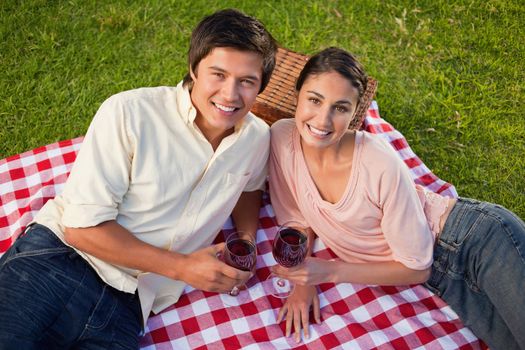 Man and a woman looking towards the sky and holding glasses of red wine while lying on a red and white picnic blanket