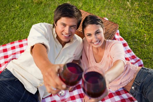 Man and a woman smiling happily as they touch their raised glasses of red wine during a picnic with focus on the glasses of wine