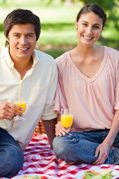 Man and a woman smiling while holding glasses of orange juice as they sit on a red and white picnic blanket