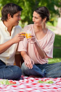 Man and a woman looking at each other while touching glasses of orange juice as they sit on a red a white picnic blanket