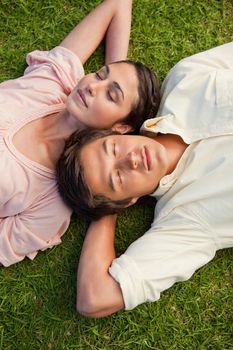 Man and a woman with their eyes closed lying head to shoulder with their arms resting behind their neck on the grass