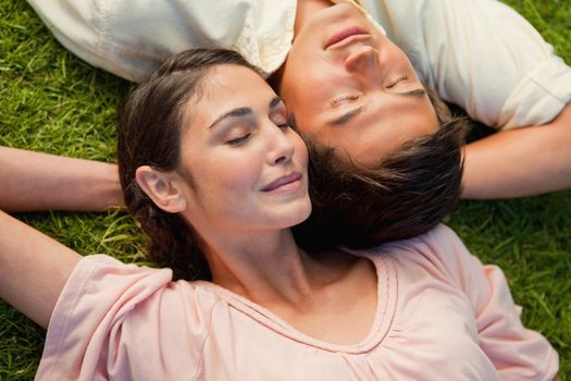 Man and a woman with their eyes closed lying head to shoulder with their arms resting behind their neck on the grass