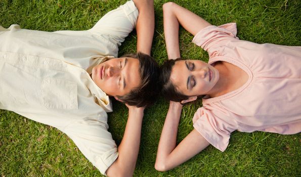 Man and a woman with their eyes closed lying head to head with both of their arms resting behind their neck on the grass