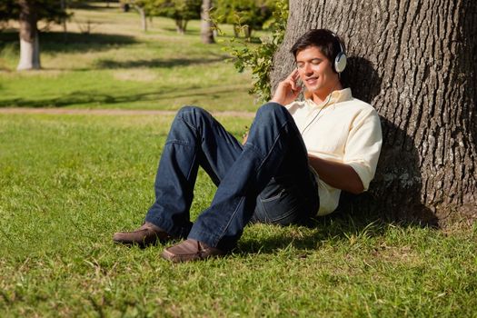 Man smiling as he uses headphones to listen to music while resting against the trunk of a tree