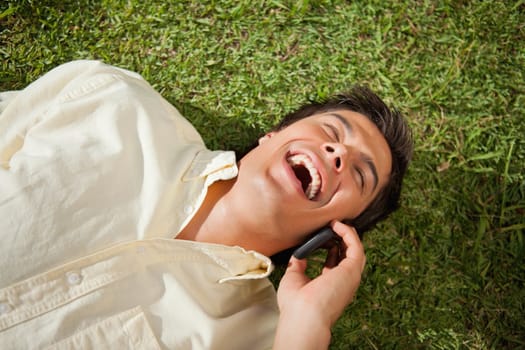 Man with his eyes closed laughing while making a call over the phone as he lies down on the grass