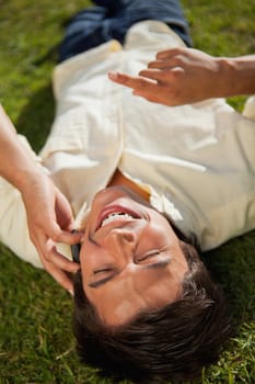 Man with his hand raised laughing while making a call over the phone as he lies down on the grass