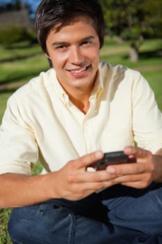Man smiling while he looks in front of him while sending a text message as he is sitting down on the grass
