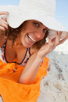 Young woman holding her hat brim while lying on a beach towel and smiling